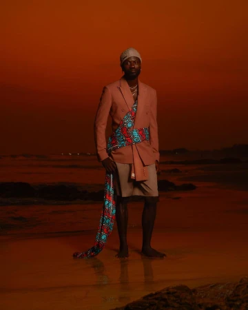 Man wearing a pink blazer, shorts, beanie, and patterned sash standing on a sandy beach at sunset.