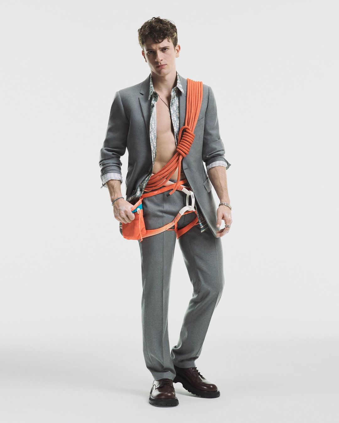 Fashion portrait of a handsome man in a grey suit with a climbing harness and rope, posing in a light studio setting.