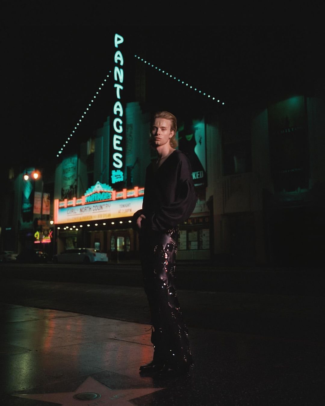 A man in dark attire stands on a wet sidewalk at night in front of the illuminated Pantages marquee, photographed by Kenny Germé.