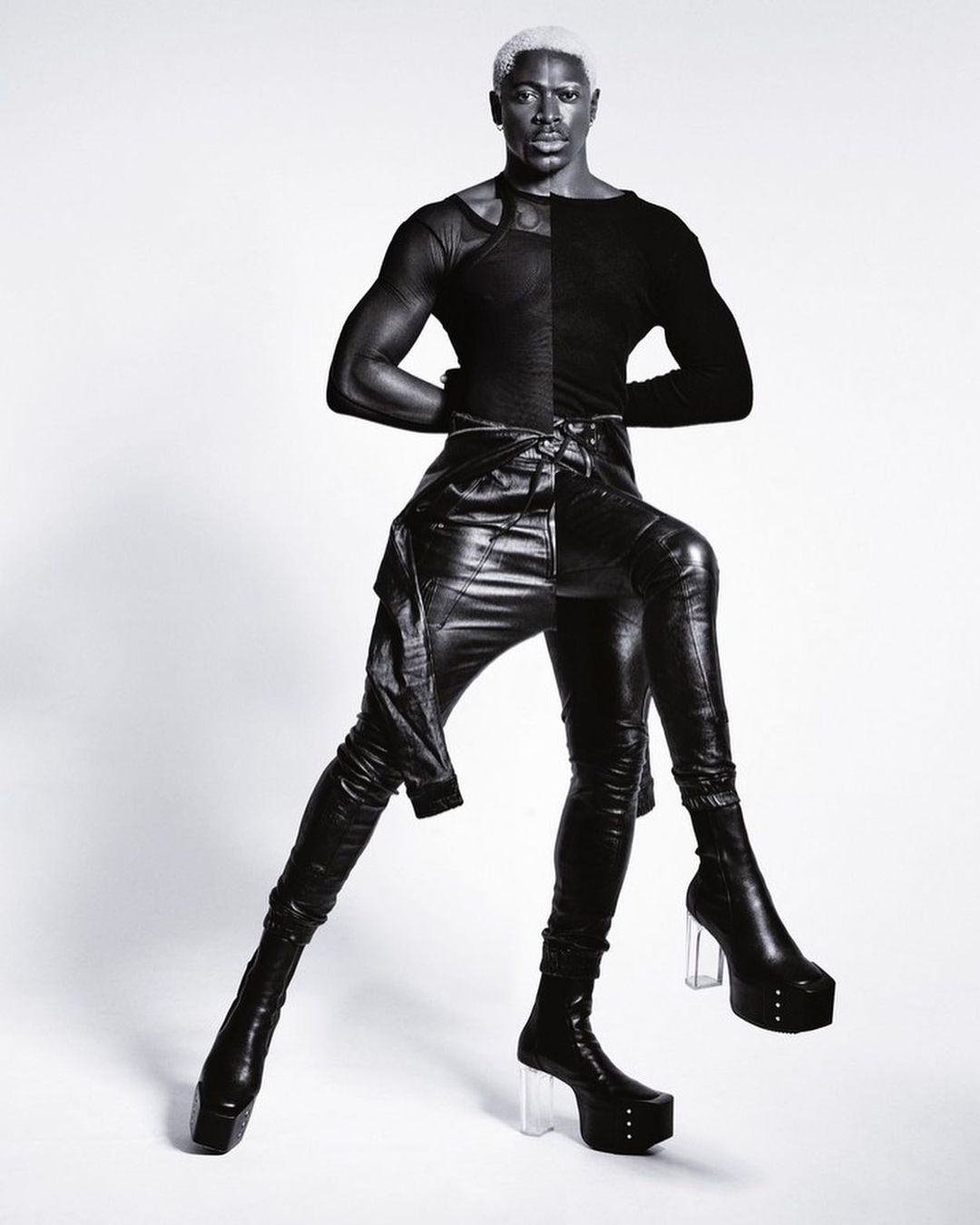 Portrait of a Black male model with bleached hair wearing black leather pants, long sleeved top, and platform boots against a white background.