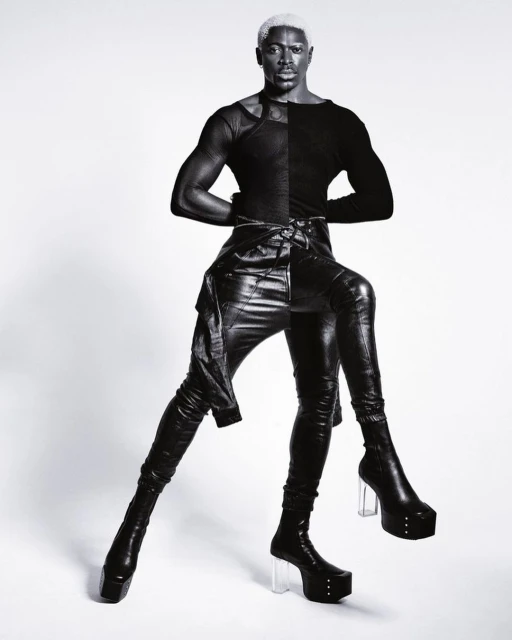 Portrait of a Black male model with bleached hair wearing black leather pants, long sleeved top, and platform boots against a white background.