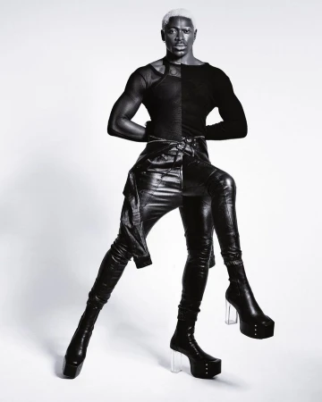 Portrait of a Black male model with bleached hair wearing black leather pants, long sleeved top, and platform boots against a white background.