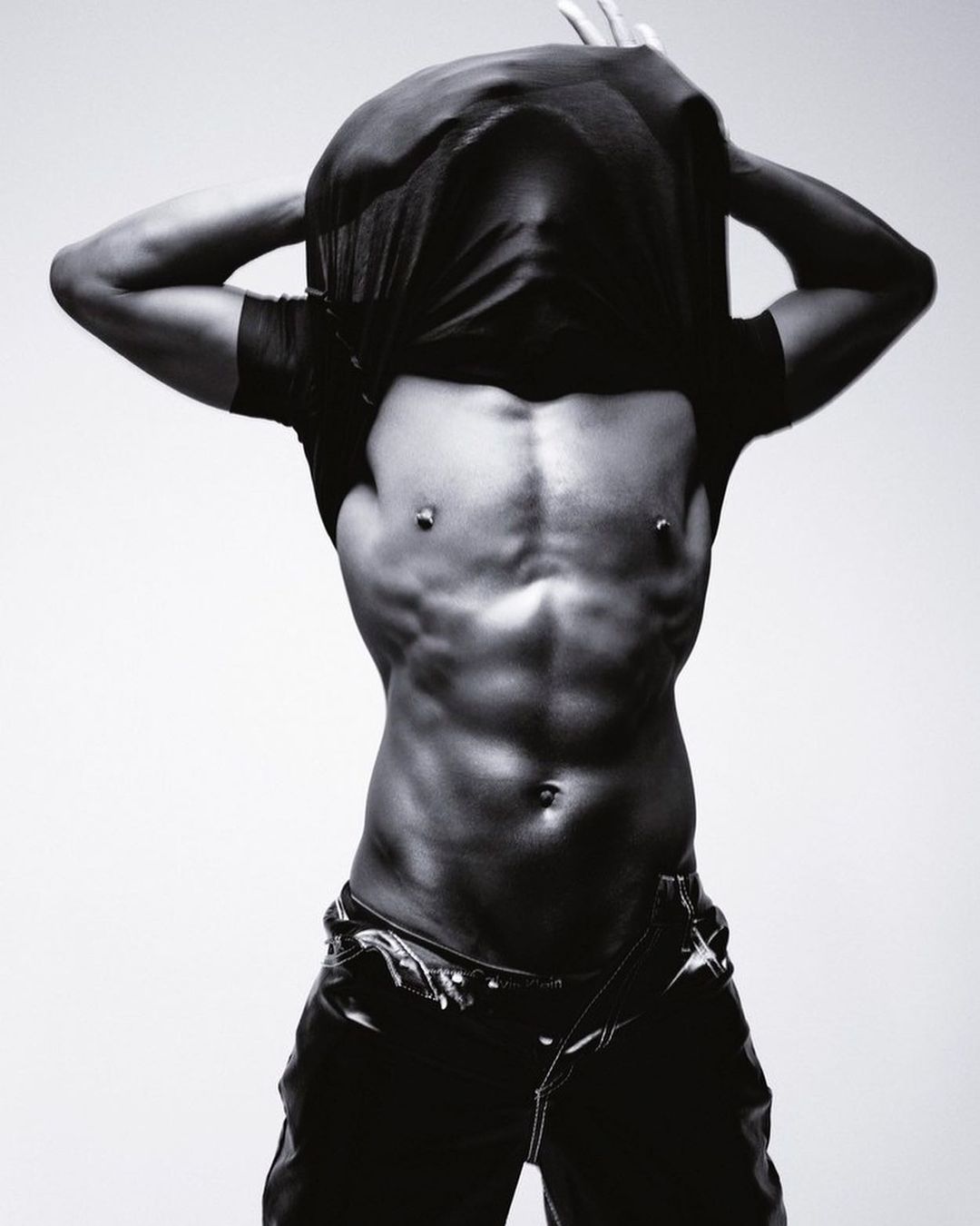 A muscular African-American male model removes his t-shirt in a black and white studio shot against a gradient grey backdrop.