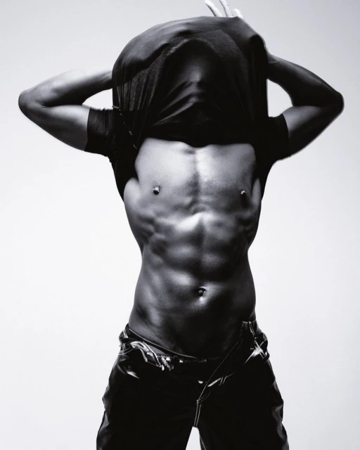 A muscular African-American male model removes his t-shirt in a black and white studio shot against a gradient grey backdrop.