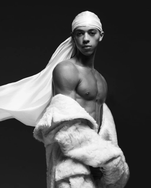 Portrait of muscular man in a white fur coat and a white durag, posing in a photo studio with a grey background in black and white.