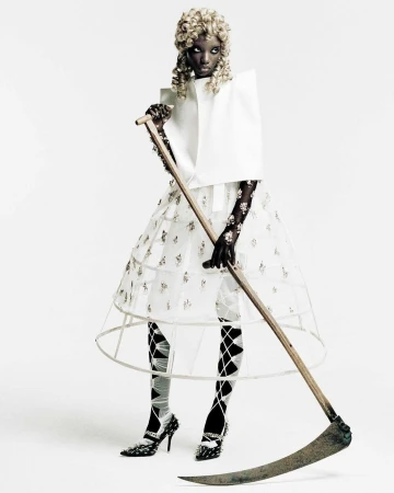 Fashion editorial shot of a Black woman wearing a white avant-garde dress and holding a scythe, posing in a bright white photo studio.