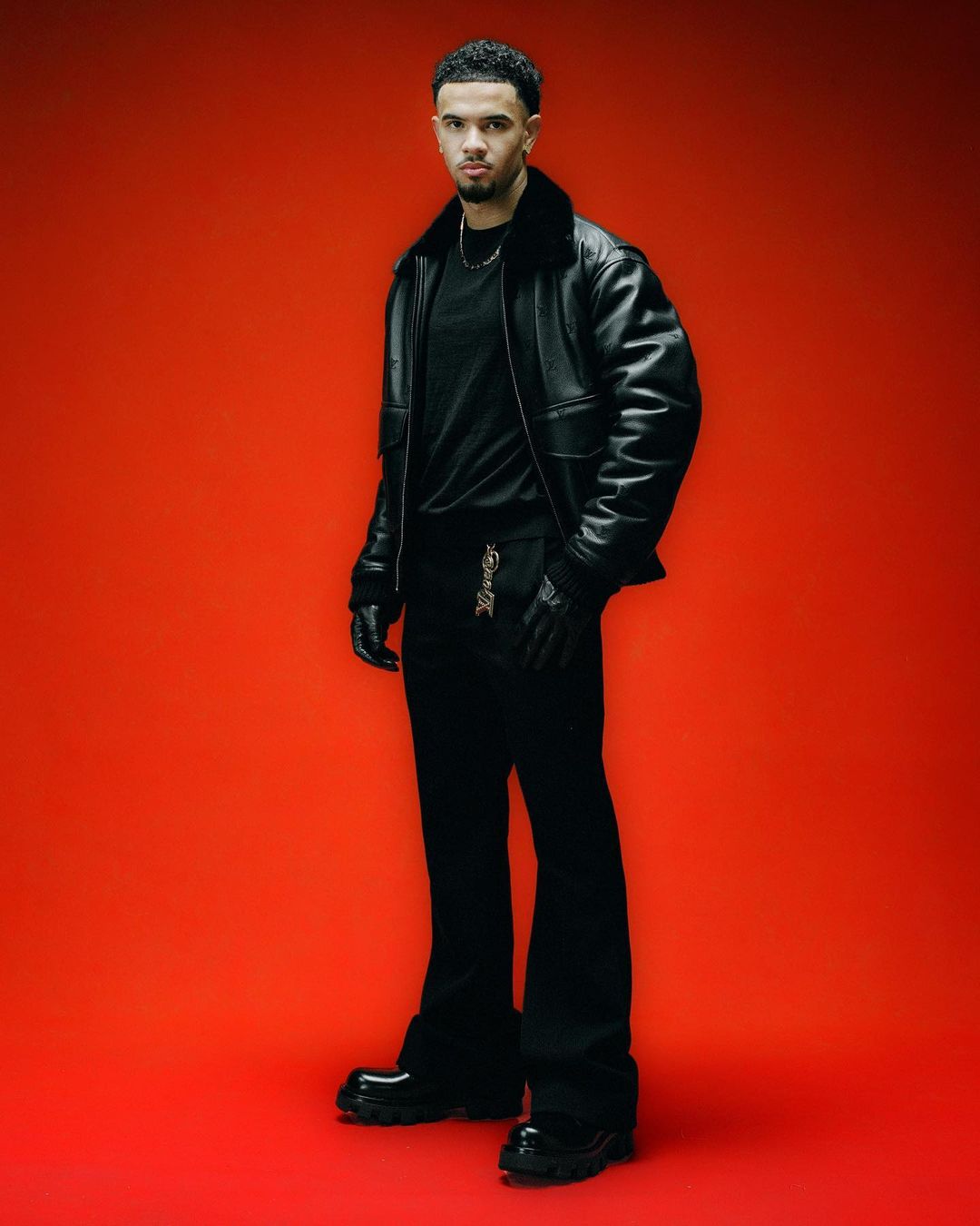 Stylish adult male in black leather jacket and pants stands against a red background in a full-length studio fashion shot.