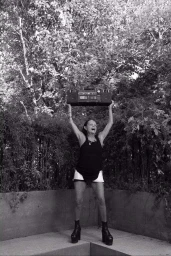 Monochrome portrait of an older woman lifting a boombox above her head in an outdoor garden setting by photographer Kenny Germé