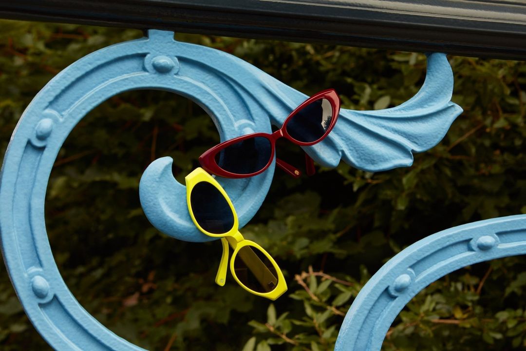 Aesthetic still life featuring red and yellow sunglasses balanced on a decorative blue metal railing, set against a blurred green backdrop.