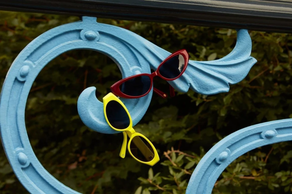 Aesthetic still life featuring red and yellow sunglasses balanced on a decorative blue metal railing, set against a blurred green backdrop.