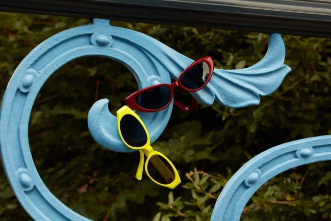 Aesthetic still life featuring red and yellow sunglasses balanced on a decorative blue metal railing, set against a blurred green backdrop.