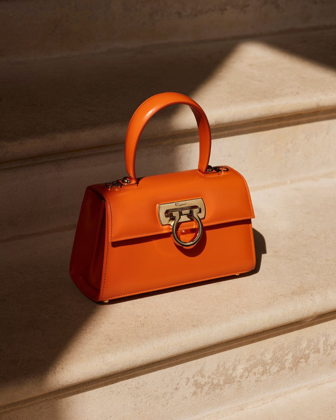 Vibrant orange leather handbag with metallic gold clasp resting on stone stairs in warm sunlight