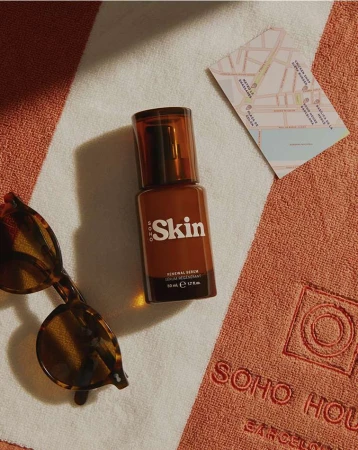 Product shot of brown glass bottle of facial serum against a white and orange striped towel with sunglasses and a map.