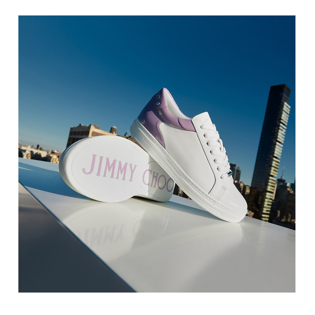 Elevated view of Jimmy Choo white leather sneakers with lilac detail on a glossy white surface with an urban skyline and bright blue sky.
