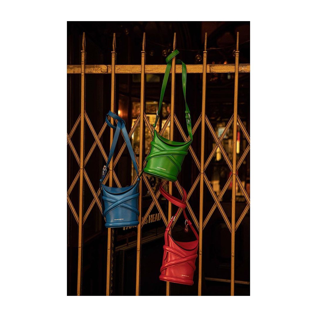 Three vibrant designer bucket bags in blue, green, and red hanging from a gold metal gate, displayed for a fashion product shot.