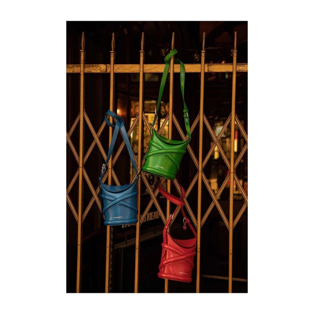 Three vibrant designer bucket bags in blue, green, and red hanging from a gold metal gate, displayed for a fashion product shot.