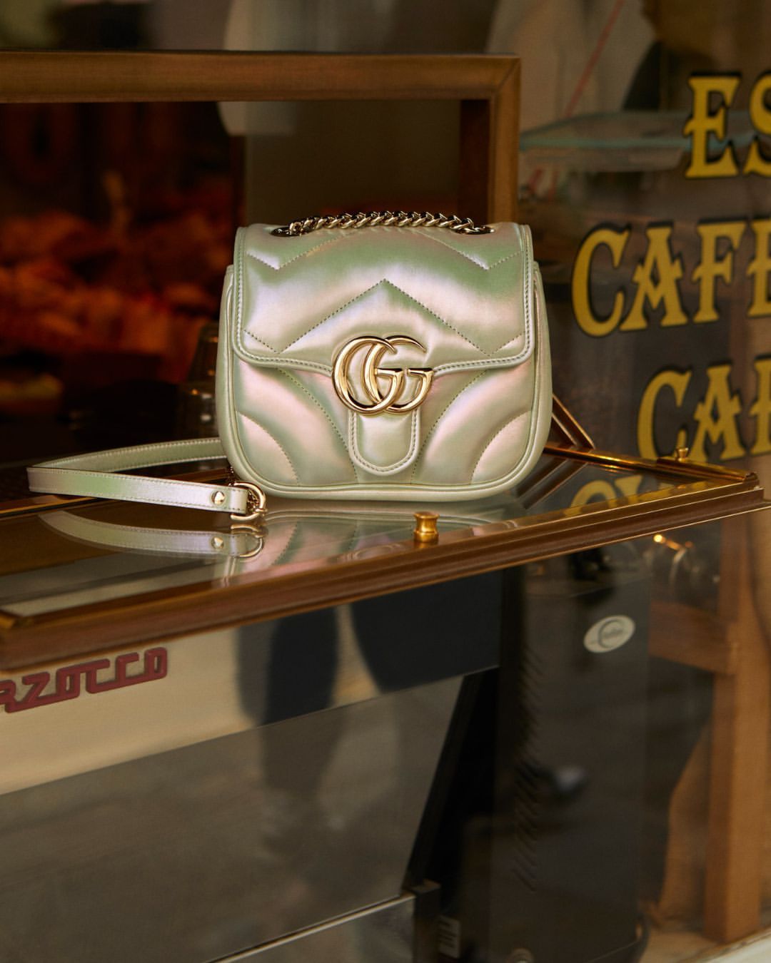 Iridescent Gucci Marmont handbag with gold hardware and chain strap displayed in front of a cafe with brown and gold signage.