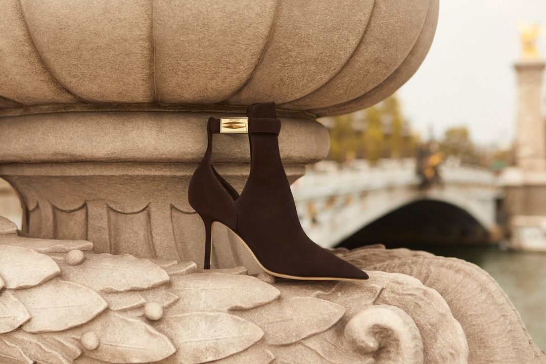 Elegant high-heeled ankle boot with gold hardware detail displayed on a stone balustrade with a bridge in the soft-focus background.