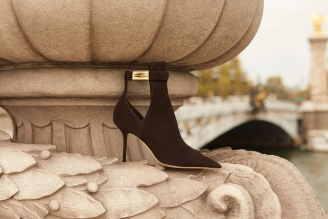 Elegant high-heeled ankle boot with gold hardware detail displayed on a stone balustrade with a bridge in the soft-focus background.