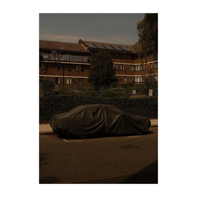 Car under a black cover on a city street with an apartment building in the background and tree casting a shadow in the street.