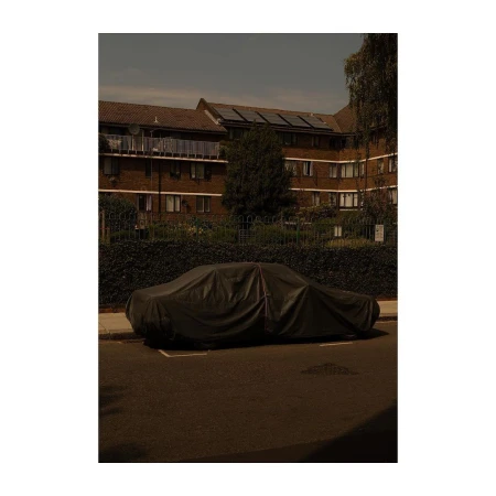 Car under a black cover on a city street with an apartment building in the background and tree casting a shadow in the street.