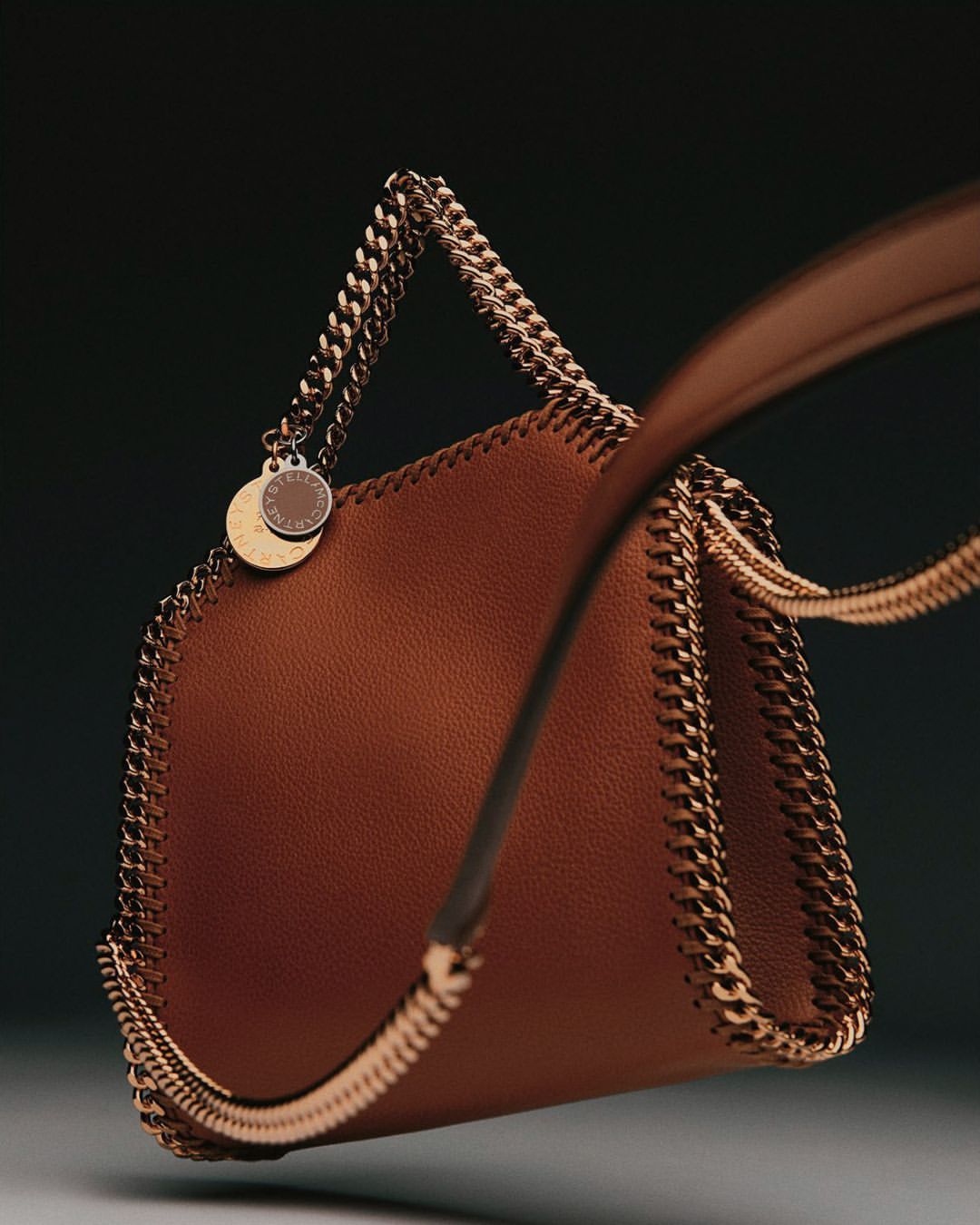 Studio shot of a brown Stella McCartney handbag with chain trim and handle, against a dark gradient background.