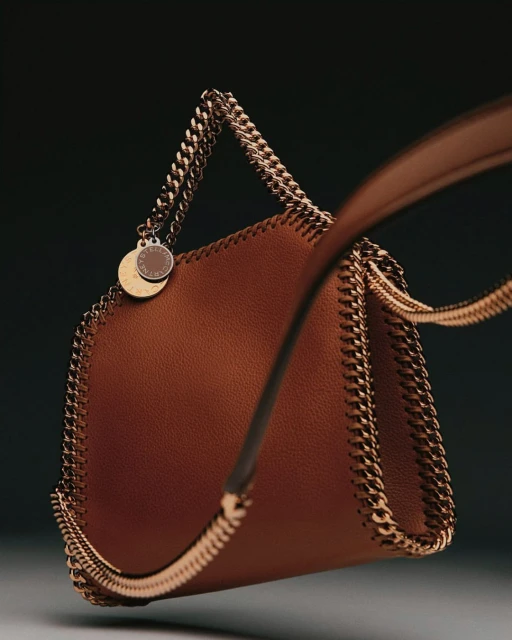Studio shot of a brown Stella McCartney handbag with chain trim and handle, against a dark gradient background.