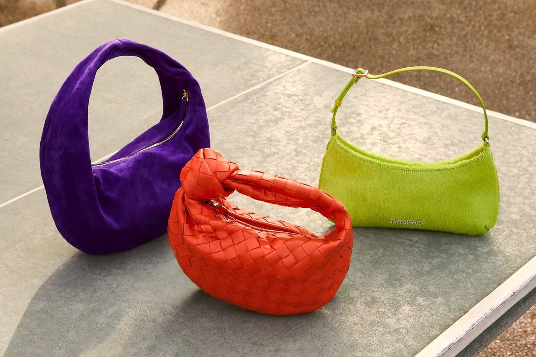 Studio shot of a bright purple, red-orange, and neon green handbags with handles. Modern fashion accessories are displayed against a gray surface.