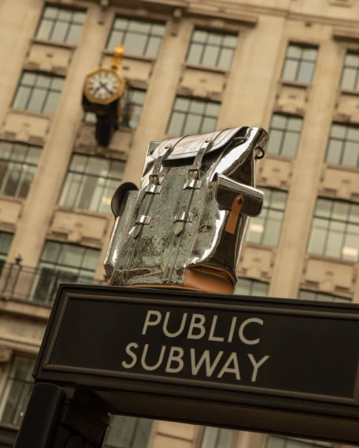 Metallic silver backpack atop a public subway entrance sign, with a clock and building facade in the background on an overcast day.