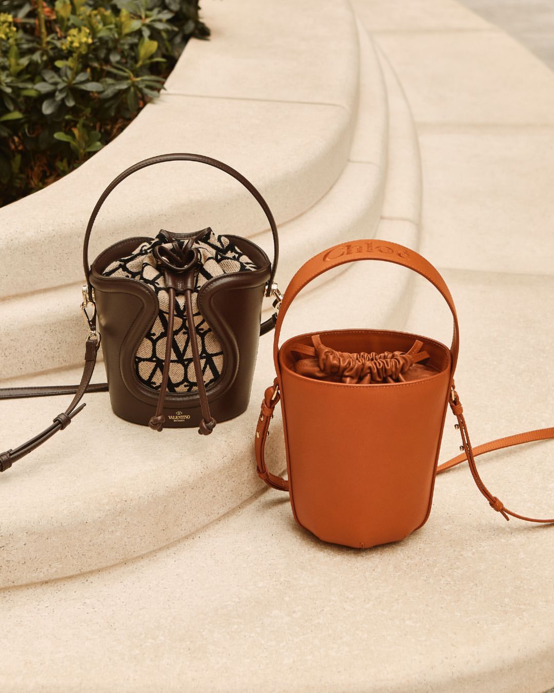 Two designer bucket bags, one brown with Valentino logo and one tan with Chloe logo, are displayed on a cream-colored stone surface.
