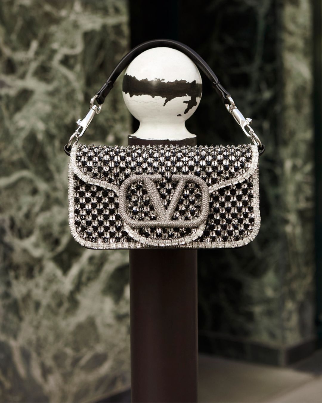 Black and silver Valentino handbag displayed on a white post with a dark background. Luxury fashion accessory with signature logo.