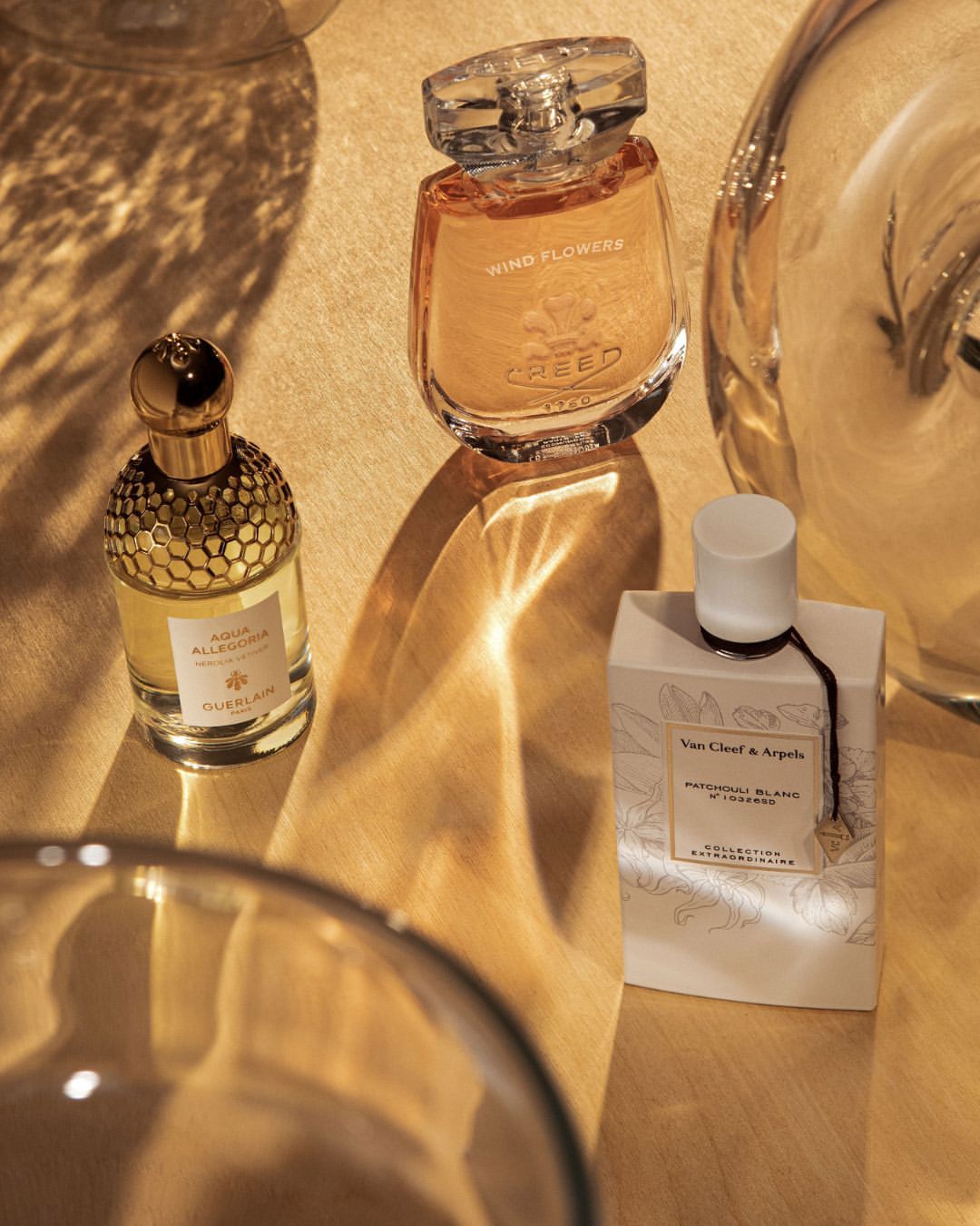 Three perfume bottles of different brands (Creed, Guerlain, and Van Cleef & Arpels) are arranged on a golden surface with light and shadow play.