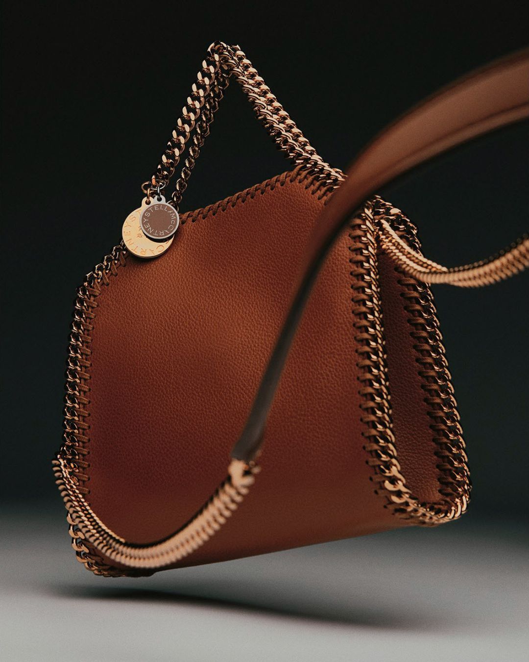 Fashion shot of a brown Stella McCartney Falabella handbag with a chain trim and strap against a dark gradient background.