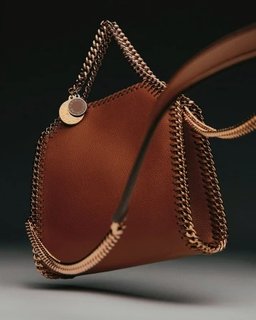 Fashion shot of a brown Stella McCartney Falabella handbag with a chain trim and strap against a dark gradient background.