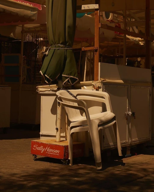 A stack of white plastic chairs sits on a red and white base with an umbrella in the background in an outdoor setting.