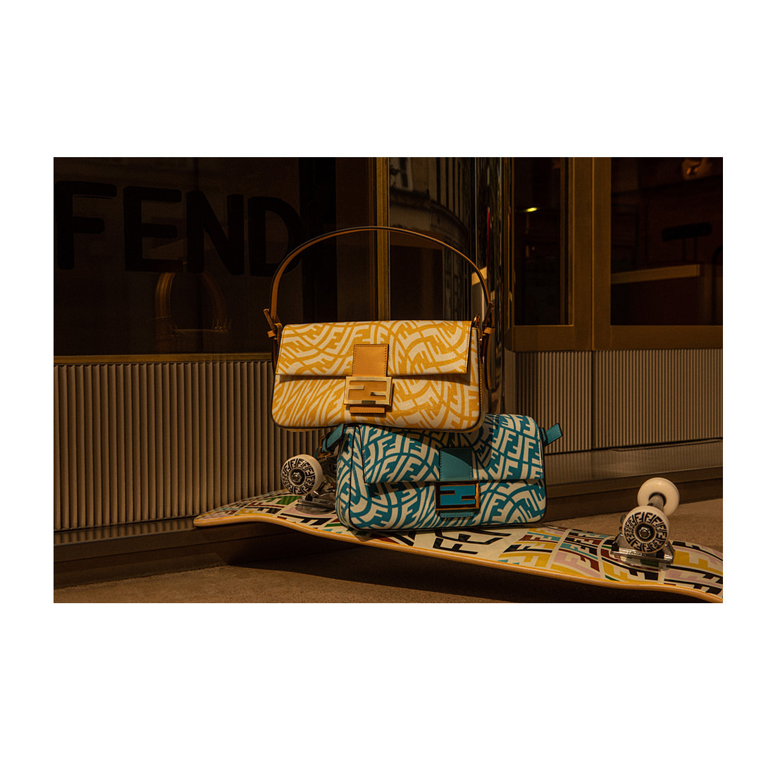 Two Fendi handbags, one yellow and white and one blue and white, are stacked on a patterned skateboard against a textured wall.