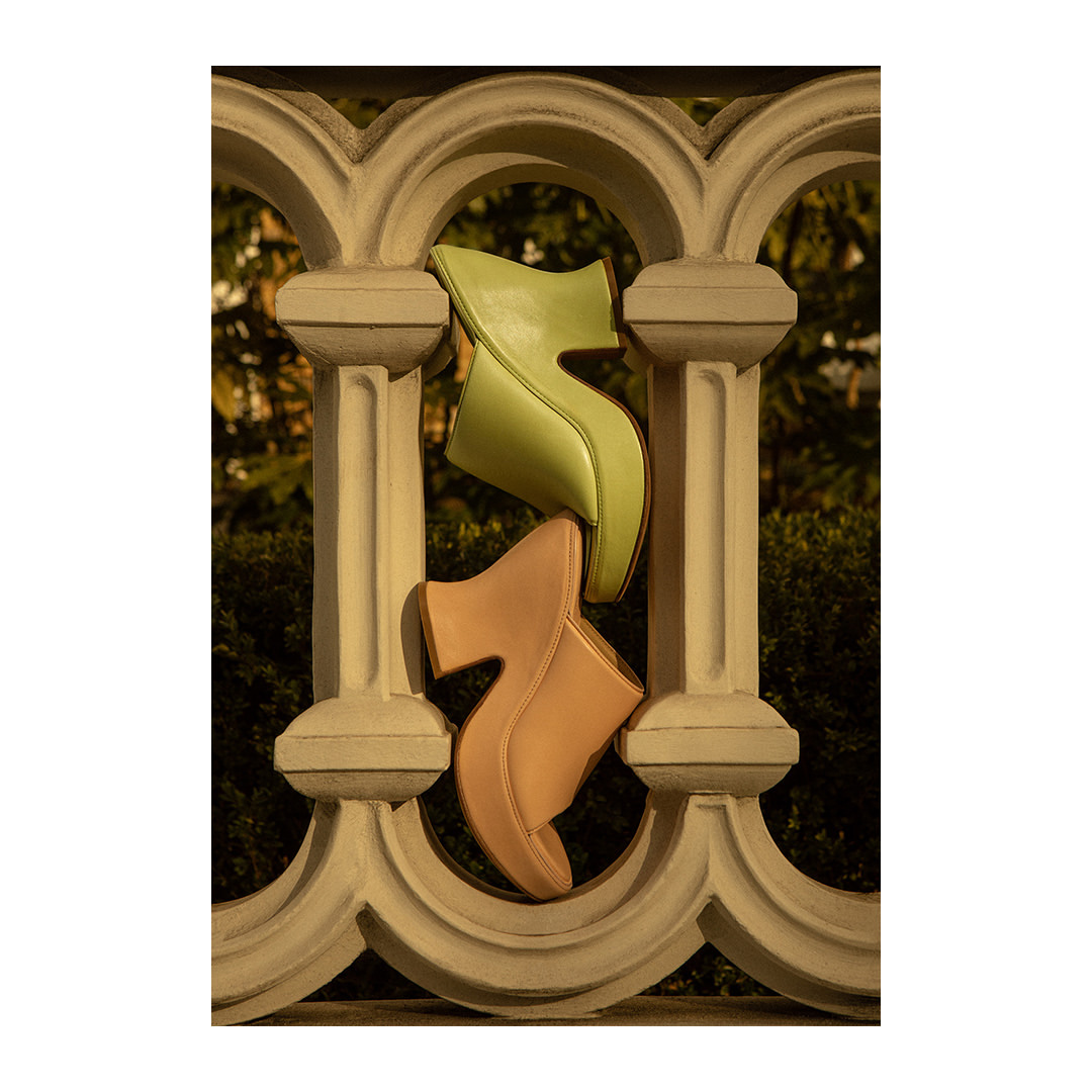 Two stylish women's platform shoes, one green and one tan, are suspended between the ornate arches of a light-grey stone balustrade.