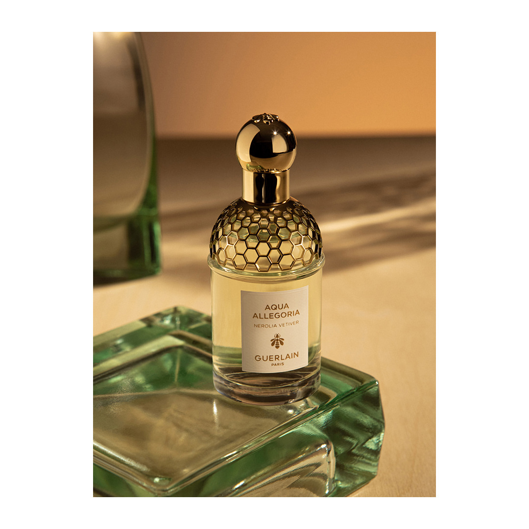 Guerlain Aqua Allegoria Nerolia Vetiver fragrance in clear bottle with gold honeycomb cap on a green glass stand against tan background.