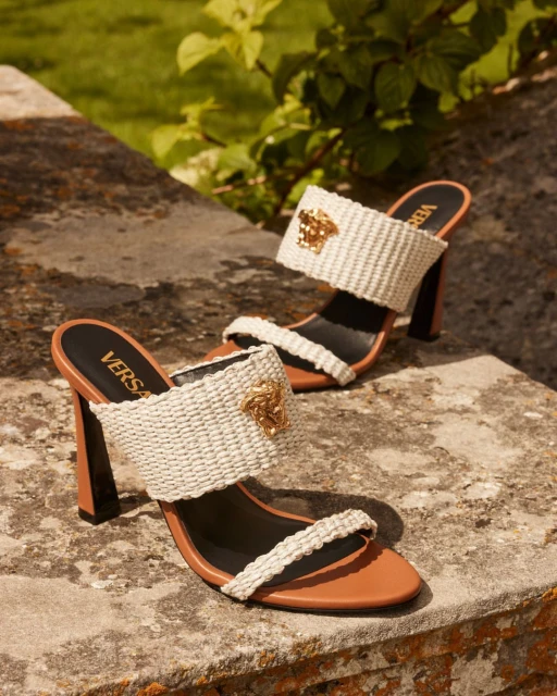 Luxury Versace branded heeled sandals in tan leather and woven raffia, featuring a gold Medusa head emblem. Outdoor setting with greenery.