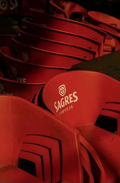 Stacked red plastic chairs with the Sagres beer logo, indoors. A collection of chairs are stacked together in a dimly lit space.