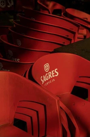 Stacked red plastic chairs with the Sagres beer logo, indoors. A collection of chairs are stacked together in a dimly lit space.