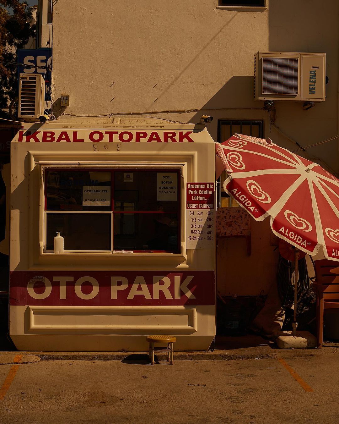 Outdoor shot of an IKBAL OTOPARK booth with a red Algida umbrella beside it, casting shadows on the sandy ground.