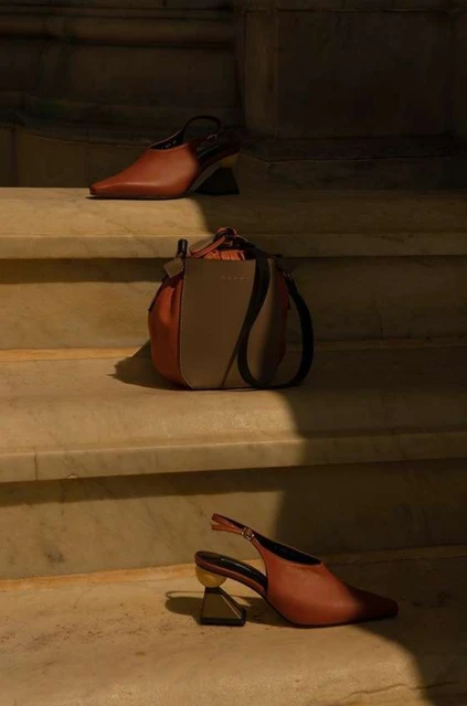 Fashion product shot: two rust-colored slingback shoes and a color-block bucket bag arranged on marble steps in warm, shadowed light.