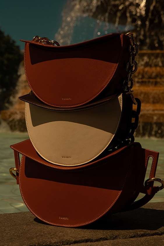 Three VAUGE bags are stacked on a stone surface with a fountain in the background, showcasing the brand's fashion accessories.