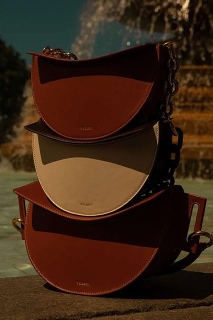 Three VAUGE bags are stacked on a stone surface with a fountain in the background, showcasing the brand's fashion accessories.