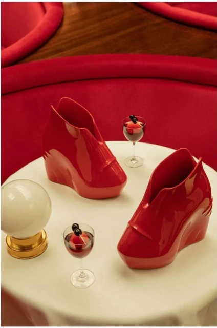 Still life composition featuring two sculptural red wedge shoes, two cocktail glasses, and a spherical lamp on a white table against a red backdrop.