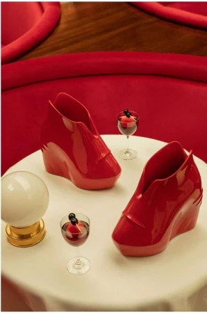 Still life composition featuring two sculptural red wedge shoes, two cocktail glasses, and a spherical lamp on a white table against a red backdrop.