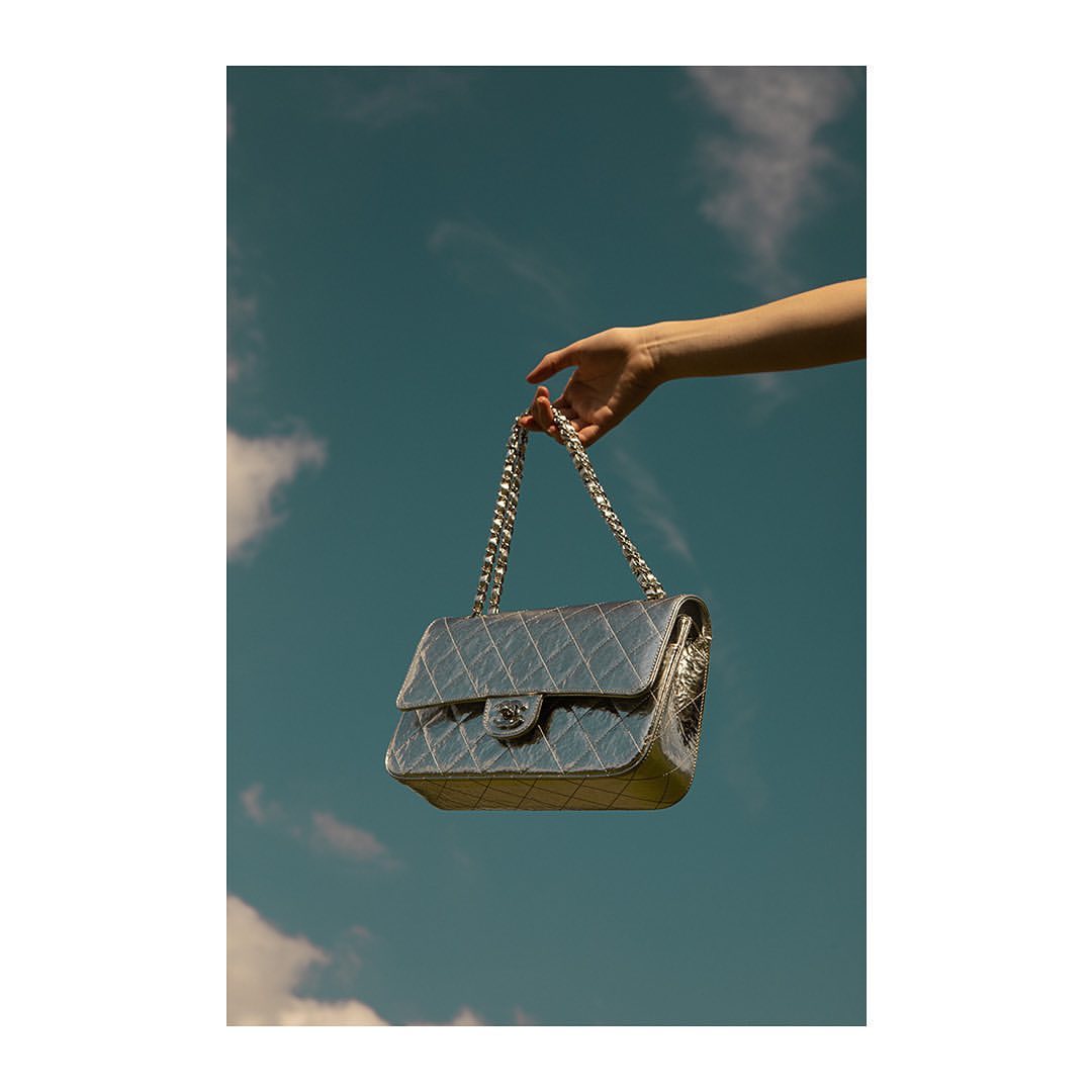 Silver Chanel handbag held by a person's hand against a backdrop of a blue sky with scattered clouds, creating a stylish and elegant visual.