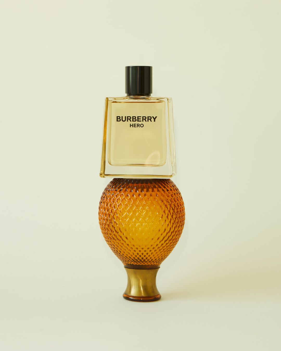 Still life shot of a Burberry Hero glass perfume bottle balanced on an orange textured glass lamp base on a light yellow background.