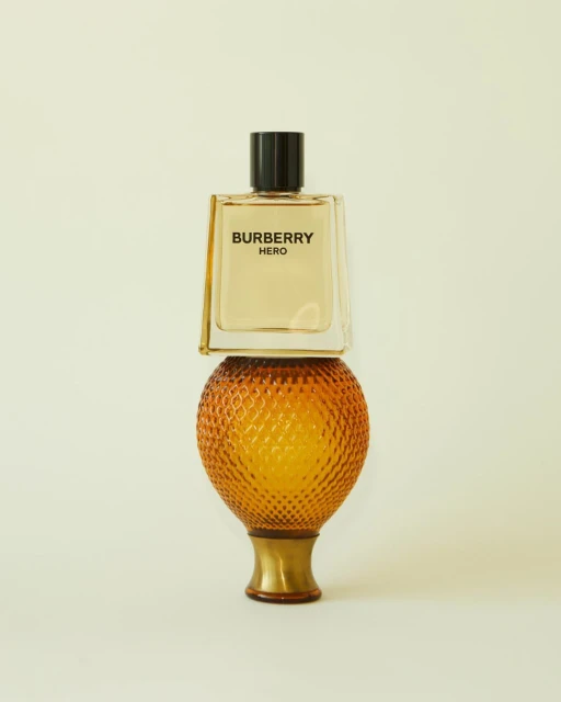 Still life shot of a Burberry Hero glass perfume bottle balanced on an orange textured glass lamp base on a light yellow background.